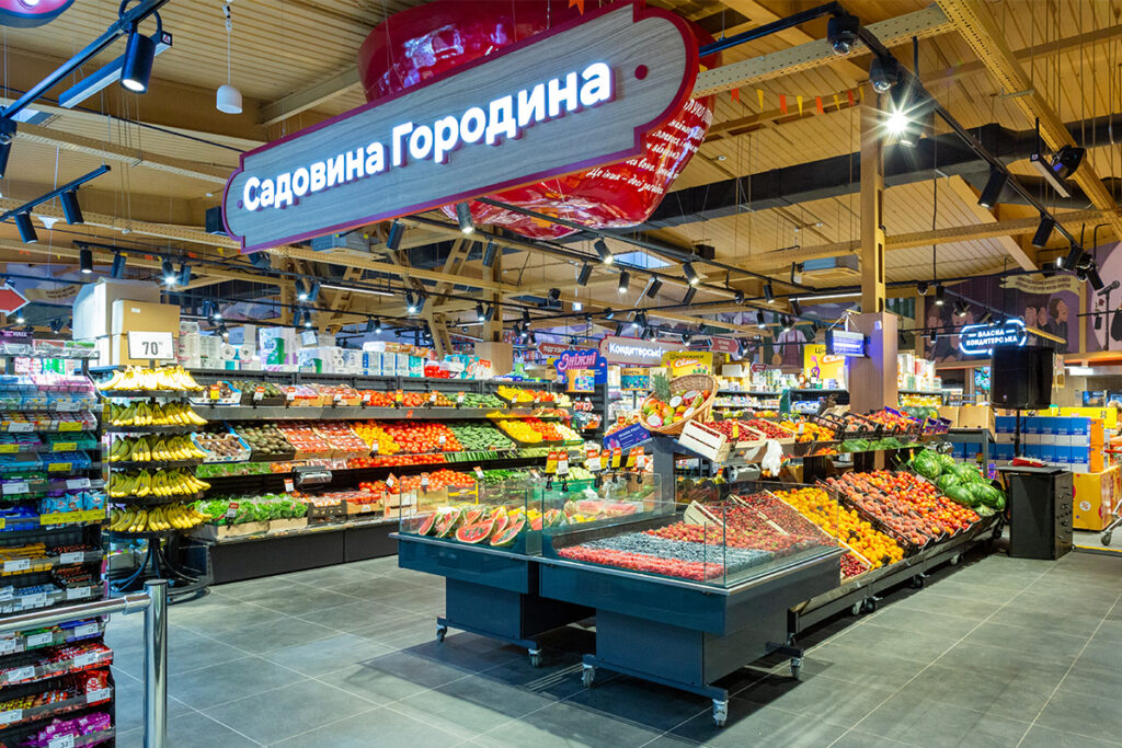 Inside a supermarket produce department with abundant fruit and vegetable displays under a large Cyrillic sign overhead.