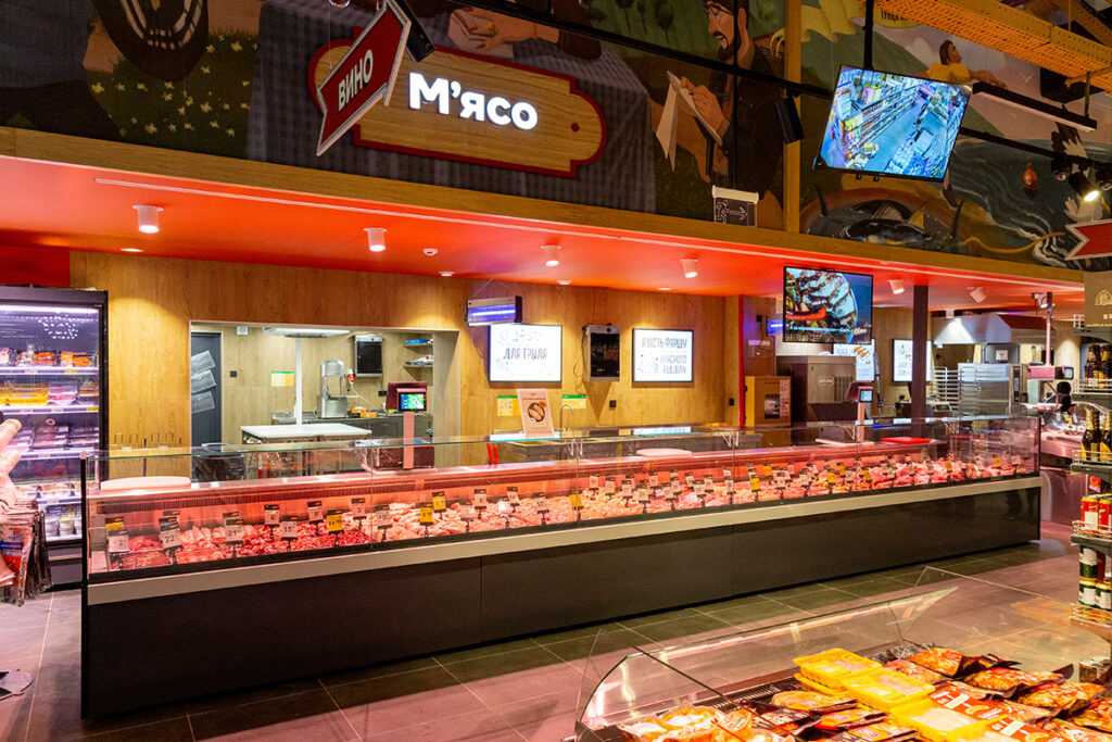 Meat counter in a grocery store with a glass display case and illuminated signs overhead (Cyrillic signage)