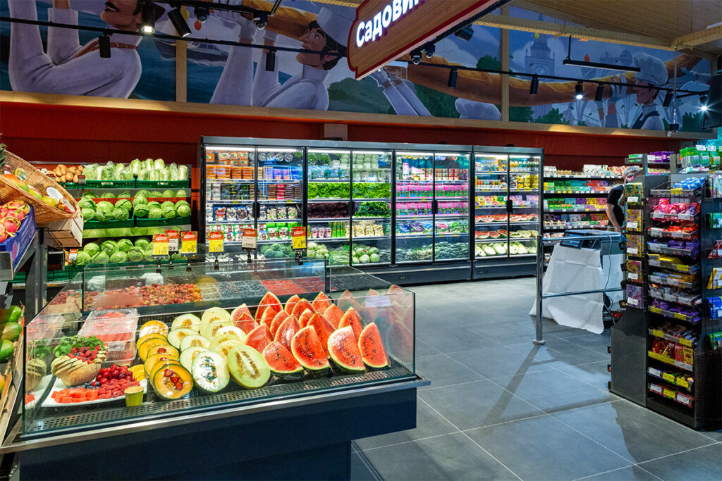 Inside a supermarket produce area with a glass fruit case, leafy greens, and refrigerated shelves; a shopper browses on the right.