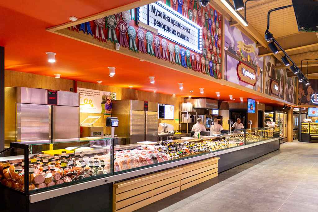 Long deli counter with glass display of meats and sausages, orange ceiling, and staff behind the counter in a bright supermarket setting
