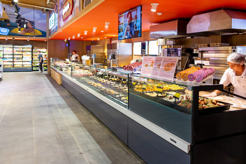 Deli counter in a supermarket with glass display cases of prepared foods and a worker in a white uniform behind the counter.