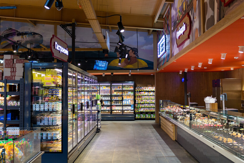 Interior of a grocery store with tall refrigerated display cases on the left and a glass deli counter along the right wall.