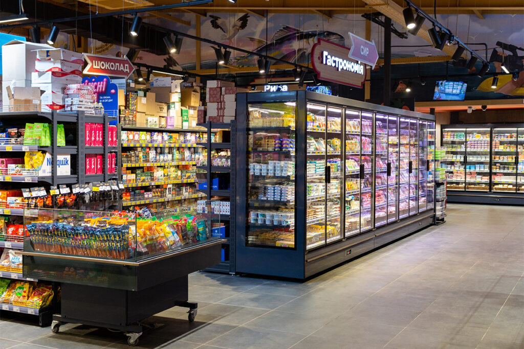 Interior of a supermarket with glass-door refrigerated units full of groceries and a snack display in the foreground.