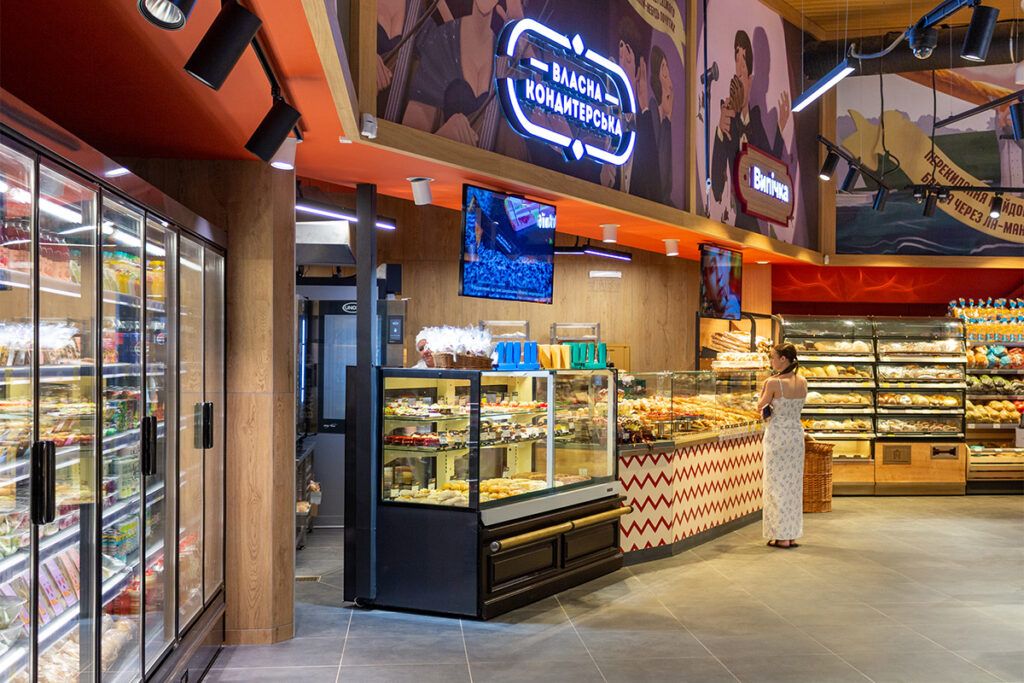 Inside a modern grocery store bakery with glass pastry displays; a woman in a white dress browses the pastries at the counter.