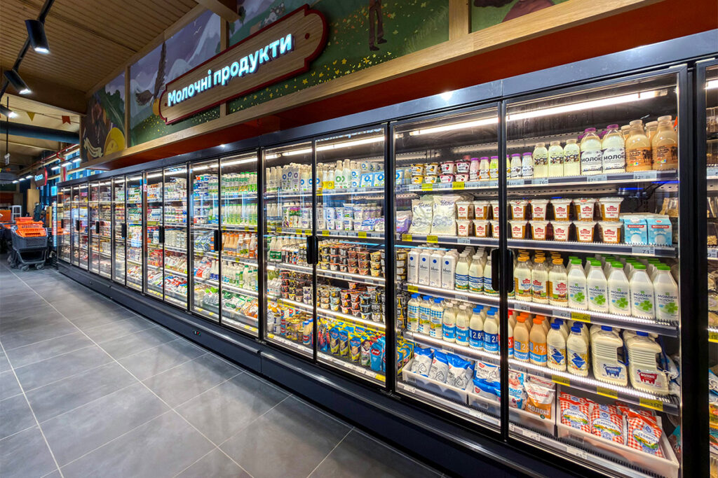 A supermarket dairy aisle with glass-door chillers filled with milk, yogurt, and other dairy products under bright lighting, extending along the aisle.